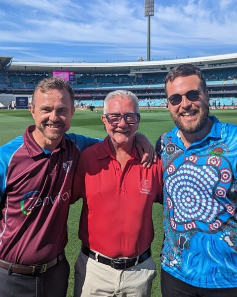 Three men standing together on a grass field at a large sports stadium with blue seating in the background