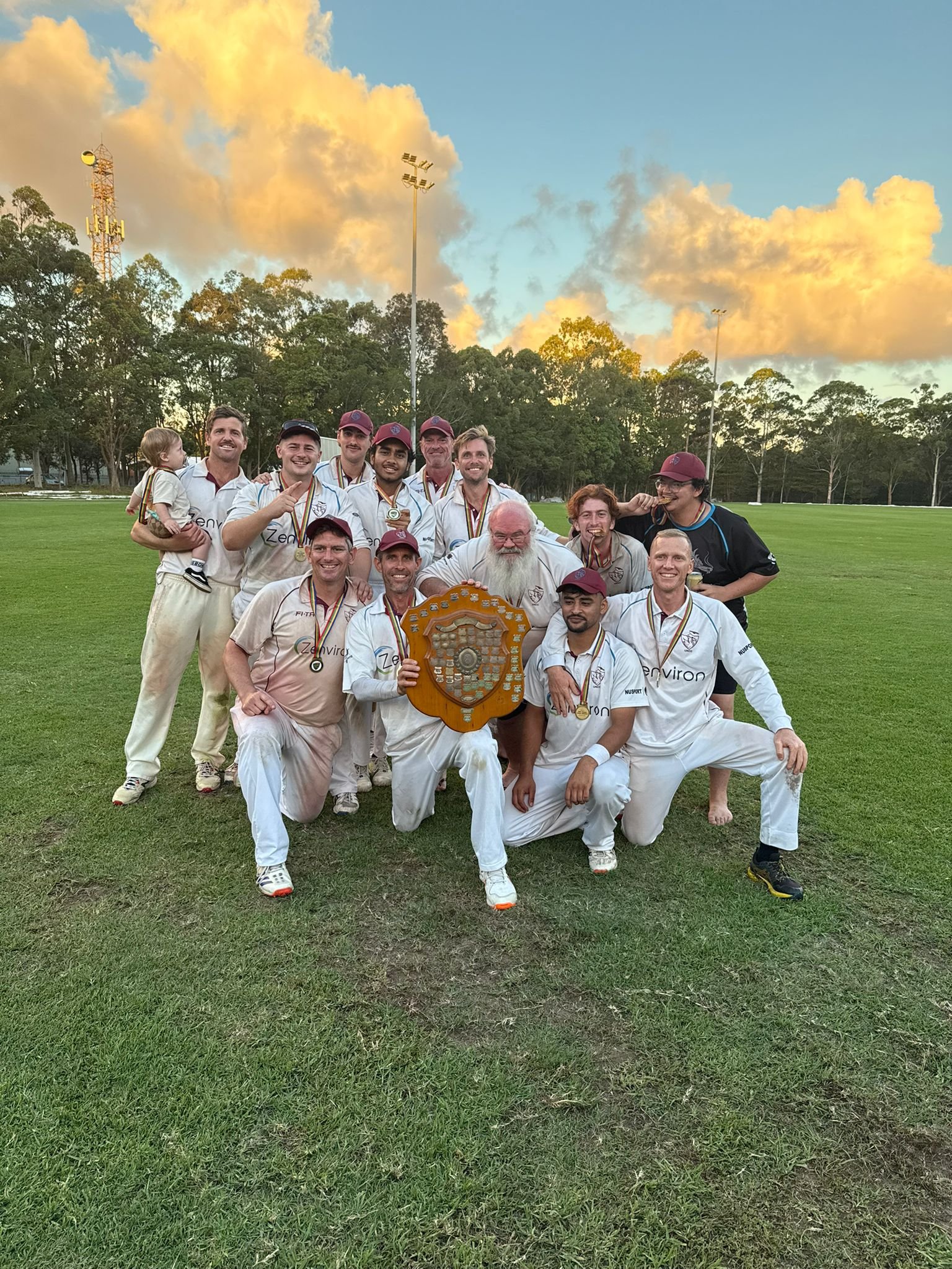 Cricket team in white uniforms posing with trophy on grass field at sunset