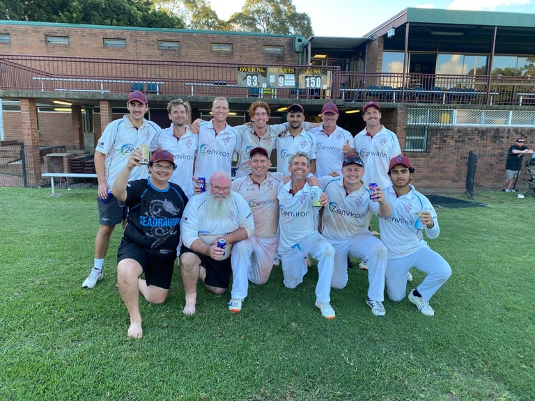 Group photo of a cricket team in white uniforms posing on grass in front of a brick stadium building