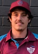 Man wearing red cap and burgundy jacket with blue shirt, standing against dark brick wall