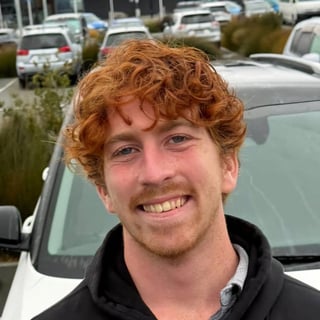 Smiling man with red curly hair and beard wearing a black hoodie, photographed outdoors with parked cars in background