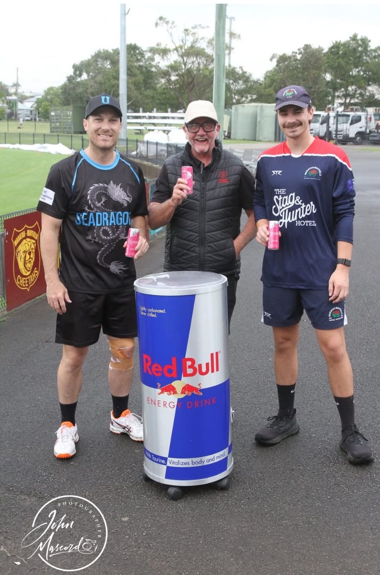 Three young men in athletic wear pose with a giant Red Bull energy drink can at a sports field, holding pink cans