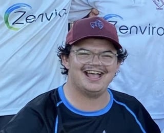 Smiling young man wearing glasses and a maroon baseball cap with blue-trimmed black shirt at an event