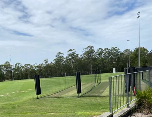 Empty sports field with metal fencing, tall light poles, and eucalyptus trees in the background under a partly cloudy sky