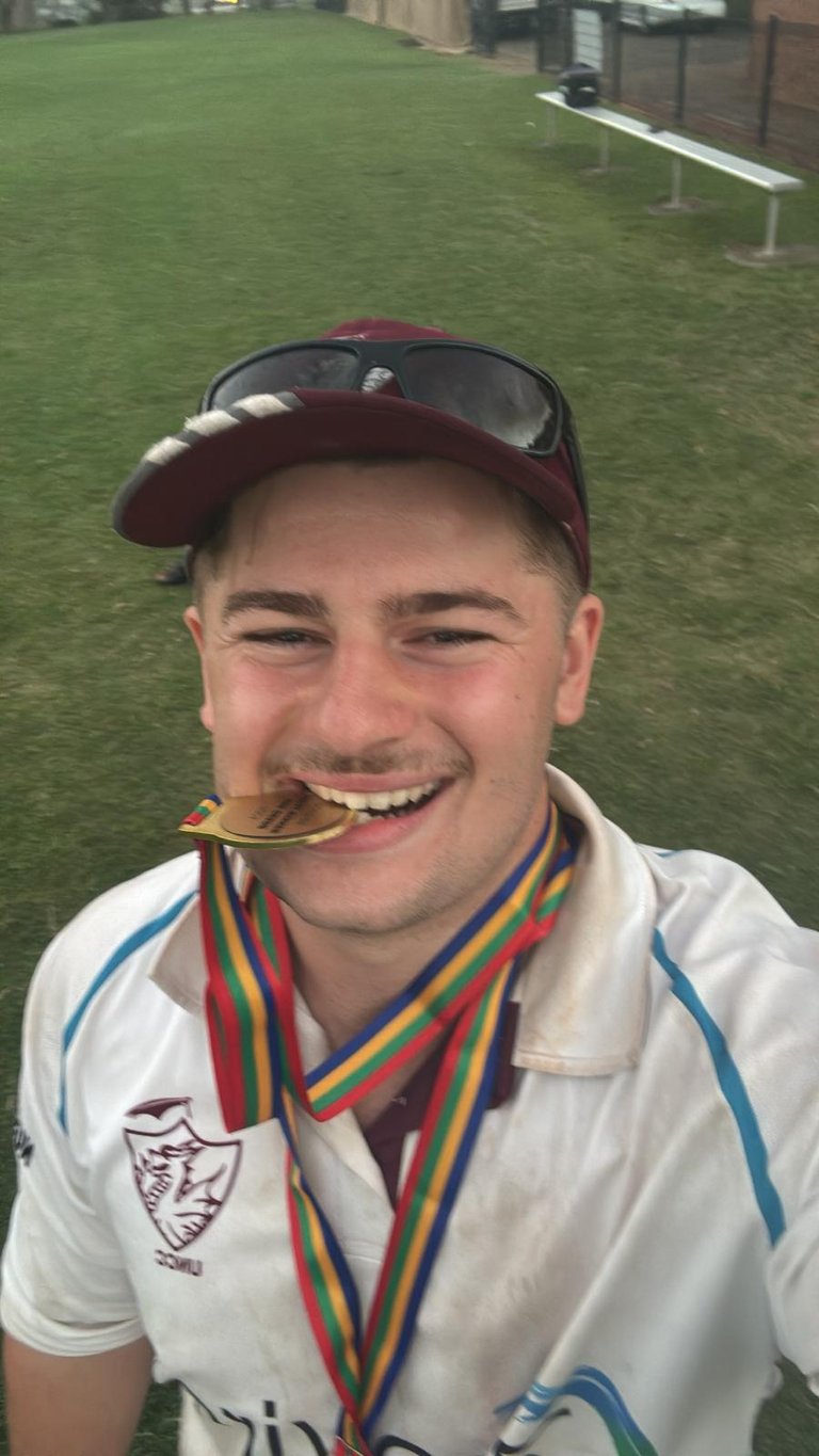 Young man in maroon cap and white sports shirt smiling while holding a gold medal with rainbow ribbon in his mouth outdoors on grass